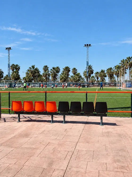 Dugout with orange and black seats beside the pitch, with palm trees behind